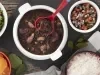 Imagem vista de cima de uma travessa redonda com feijoada, ao lado um pote de vinagrete, um pote de arroz, um pote de laranja e um pote de couve.