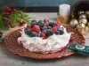 Foto da receita de pavlova natalina, branca, sobre um prato vermelho, decorada com frutas vermelhas e açúcar. Ela está numa mesa de madeira preenchida de decorações de natal como pinhas, bolas de natal e uma vela acesa ao fundo