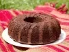 Fotografia de um bolo de chocolate em forma de bundt, com uma cor marrom profunda e textura densa, colocado em um prato branco. O fundo é um tecido listrado em vermelho e verde.