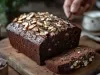 Bolo de chocolate denso, parcialmente fatiado, com amêndoas fatiadas por cima. Ele repousa em uma tábua de madeira, com uma mão segurando uma faca ao fundo, criando uma atmosfera acolhedora e convidativa.