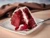Fotografia de uma fatia de bolo veludo vermelho em um prato cerâmico branco, com creme de queijo, e um garfo prateado ao lado. O fundo é um tecido marrom suave, sobre uma mesa de madeira clara.