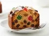 Fotografia de uma fatia de bolo de frutas em um prato branco, com uma crosta dourado-marrom e recheada com frutas cristalizadas, passas, decorado com amêndoas e polvilhada com açúcar de confeiteiro.