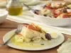 Imagem de um prato branco com uma porção de bacalhoada com ovos, pimentões, azeitona preta, tomate e cebola com um garfo ao lado. Ao fundo, uma travessa de bacalhoada e um vidro de azeite.