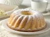 Um bolo tipo bundt (em formato de anel) com uma casquinha dourada e cobertura de açúcar de confeiteiro. Está servido em um prato escuro ao lado de uma xícara de café, criando um clima acolhedor e caseiro.