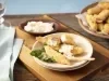 Imagem de um prato com iscas de tilápia frita e molho. Ao fundo, um bowl com iscas de tilápia e um potinho com molho.