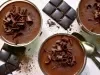 Fotografia vista de cima, em tons claros, de três recipientes de vidro sobre uma mesa clara. Dentro dos potinhos tem a sobremesa, finalizada com ganache e raspas de chocolate, e ao redor tem raspas e algumas barras pequenas de chocolate.