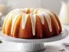 Fotografia de um bolo bundt em um suporte branco. O bolo, com textura úmida e dourada, é coberto por uma cobertura branca espesso que escorre elegantemente.