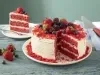 Fotografia em tons de vermelho em uma bancada de madeira clara, um prato redondo branco com o bolo red velvet cortado ao meio. Ao fundo, um prato pequeno com o pedaço.