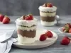 Fotografia em tons de vermelho em uma bancada de madeira clara, um paninho bege, um copo de vidro com a sobremesa de creme branco e chocolate dentro dele e em cima de um prato branco redondo. Ao fundo, mais um copo e morangos para decorar.