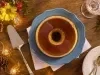 Imagem vista de cima de um pudim de leite condensado com calda de caramelo em um prato azul. Ao lado, decoração festiva e flores.