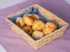 Fotografia de uma cestinha de uma cestinha de madeira retangular sobre um pano quadrado roxo. Dentro da cestinha tem um paninho menor azul com os pães de queijo.