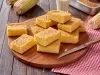 Imagem de uma tábua redonda com pedaços de bolo de fubá cremoso e uma faca em cima. Ao lado, um pano xadrez vermelho e espigas de milho.