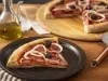 Imagem de um prato preto com uma fatia de pizza de calabresa com cebola com uma tábua de madeira e a pizza ao fundo.