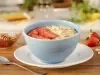 Imagem de um bowl azul claro com mingau de aveia, rodelas de morango, lascas de amêndoas e mel. Ao redor temos morangos, uma colher e um potinho de mel.