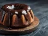 Fotografia de um bolo bundt com crosta dourada, coberto por um glaze de chocolate escuro, posicionado em uma tábua de madeira rústica.