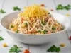 Fotografia de uma salada em uma tigela branca, composta por frango desfiado, ervilhas, milho e cenouras, coberta com maionese e finalizada com batata-palha crocantes, disposta sobre uma superfície de madeira clara.