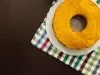 Um bolo de cenoura inteiro, redondo e simples, está colocado sobre um prato branco. A base do prato está sobre um pano de prato colorido xadrez, e o fundo é uma mesa escura de madeira, criando um contraste bonito com o bolo dourado.