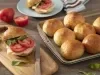Fotografia em tons de marrom em uma bancada de madeira escura, uma tábua de madeira com um sanduíche de pão de batata doce recheado com presunto, queijo e rodela de tomate. Ao lado, uma forma com vários pãezinhos.