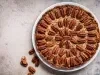 Fotografia vista de cima de uma torta inteira de nozes e doce de leite, apoiada em um prato grande, raso e branco sobre uma mesa tipo mármore. Ao lado do prato tem seis nozes sobre a mesa.