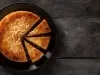Um bolo de mandioca caramelizado em formato redondo está servido em um prato preto. Algumas fatias já foram cortadas, revelando sua textura cremosa. A cobertura tem um tom dourado e está decorada com coco tostado no centro.