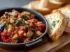 Fotografia mostra uma caponata natalina colorida servida em um prato de ferro, com cubos de berinjela dourada, pimentão e fatias de pão.