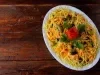Fotografia de uma salada apresentada em um prato oval branco, com ingredientes finamente picados em tons amarelos e laranjas, azeitonas verdes, batata palha e um enfeite de tomate em forma de rosa, rodeado por folhas de salsa fresca.