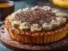 Em cima de uma mesa, banoffe com massa crocante, decorada com bananas em rodelas e canela em pó.