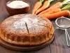 Imagem apresenta um bolo de cenoura em uma tábua de madeira, com uma crosta dourada e uma fatia cortada, revelando seu interior úmido. Cenouras frescas e uma tigela de farinha estão ao fundo, destacando a natureza caseira e apetitosa do bolo.
