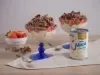 Foto da receita de pavê de leite moça com morangos e chocolate, servidos em taças com uma base azul, sobre uma bancada de madeira decorada com um tecido, pedaços de chocolate, um pote com morangos e uma lata de Leite Moça