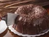 Imagem de um bolo redondo de chocolate com bastante cobertura de brigadeiro e uma mão com uma espátula cortando o bolo.
