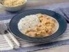 Fotografia em tons de azul em uma bancada de madeira com um jogo americano azul com listras brancas, um prato fundo azul escuro com o strogonoff de frango e arroz branco. Ao fundo, um potinho com batata palha.