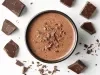 Fotografia vista de cima de uma forma funda, preta, grande e redonda sobre uma mesa branca. Dentro tem a sobremesa, finaliada com a ganache e raspas de chocolate. Ao redor tem pedaços de chocolate.