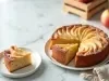 Imagem de um bolo redondo de maçã com fatias de maçã decorando sobre um prato branco. Ao lado, um prato com uma fatia de bolo e ao fundo, maçãs.
