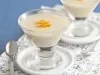 Fotografia em tons de azul em uma bancada de madeira azul, uma toalha branca com flores verdes e azuis, dois pratinhos brancos com taças de vidro com a mousse de coco e decorado com raspas de laranja.