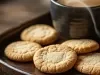 Em cima de uma mesa, prato com biscoitos redondos dourados e crocantes. Ao lado, xícara com café.