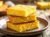 Fotografia de um empilhamento de três pedaços de bolo de fubá em um prato cerâmico rústico. A cor dourada e a textura úmida e fofa são evidentes, com um fundo que sugere uma maior quantidade do prato.