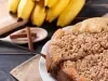Fotografia uma cuca de banana com farofa sobre um prato de vidro branco raso, apoiado em um pano marrom de tom claro. Tem uma fatia da torta sendo retirada com uma espátula de prata. Ao lado tem uma colher branca com pau de canela e bananas inteiras.