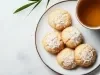 Em cima de uma mesa, prato redondo com biscoitos amanteigados, decorado com coco ralado. Ao lado direito, xícara de chá.