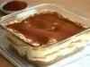 Em cima de uma mesa, refratário com porção de tiramisu com creme branco cremoso, polvilhado com cacau em pó.