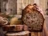 Fotografia de um bolo de nozes rústico, parcialmente fatiado, exibindo sua textura densa e úmida, com pedaços de nozes e passas. O bolo está sobre uma tábua de madeira, em um ambiente acolhedor e natural.