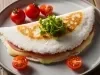 Fotografia em tons claros de uma tapioca recheada com queijo, presunto, orégano e alguns tomatinhos em volta, dentro de um prato cinza de vidro raso.