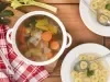 Imagem vista de cima de uma panela com caldo de legumes com pedaços de cebola, cenoura. alho poró e ao lado dois pratos de massas.