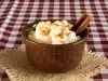 Fotografia de um recipiente pequeno de vidro marrom com uma receita de canjica vegana. O recipiente está sobre um pano de mesa quadriculado de tom vinho e branco, ao lado do recipiente tem três paus de canela.