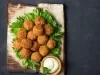 Fotografia vista de cima de várias unidades de falafel sobre uma folha de alface e um apoio de madeira retangular, ao lado um molho de iogurte que também está sobre o apoio.