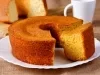 Fotografia de um bolo de milho recém-assado, com uma fatia cortada que revela seu interior macio. O bolo, em um prato com borda ondulada, com uma caneca ao fundo, transmitindo uma atmosfera caseira.