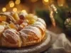 Fotografia de um bolo tradicional de frutas, embelezado com frutas cristalizadas e polvilhado com açúcar de confeiteiro. O fundo desfocado com luzes douradas e ramos de pinheiro sugere um ambiente festivo.