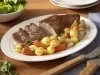 Fotografia em tons de verde em uma bancada de madeira clara, um prato oval branco com a maminha fatiada e legumes em cima dele. Ao fundo, um recipiente com salada, talheres de churrasco.