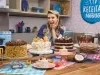 Foto da confeiteira e influenciadora Beca Milano atrás de uma bancada de cozinha repleta de bolos decorados de diversos tipos