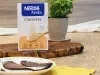 Fotografia em tons de marrom com um prato redondo ao centro. Em cima do prato existe duas panquecas de chocolate com aveia recheadas com um creme de iogurte. Ao fundo existe uma embalagem de Farinha de aveia NESTLÉ