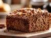 Fotografia de um pedaço de bolo denso e escuro, coberto com uma camada de chocolate ou caramelo, com pedaços visíveis de nozes ou frutas, em um cenário aconchegante com paus de canela.